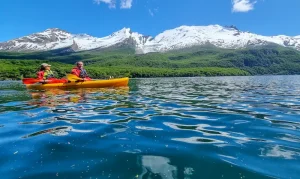 Dos personas en kayak amarillo sobre aguas azules con montanas nevadas y bosque en el Lago del Desierto